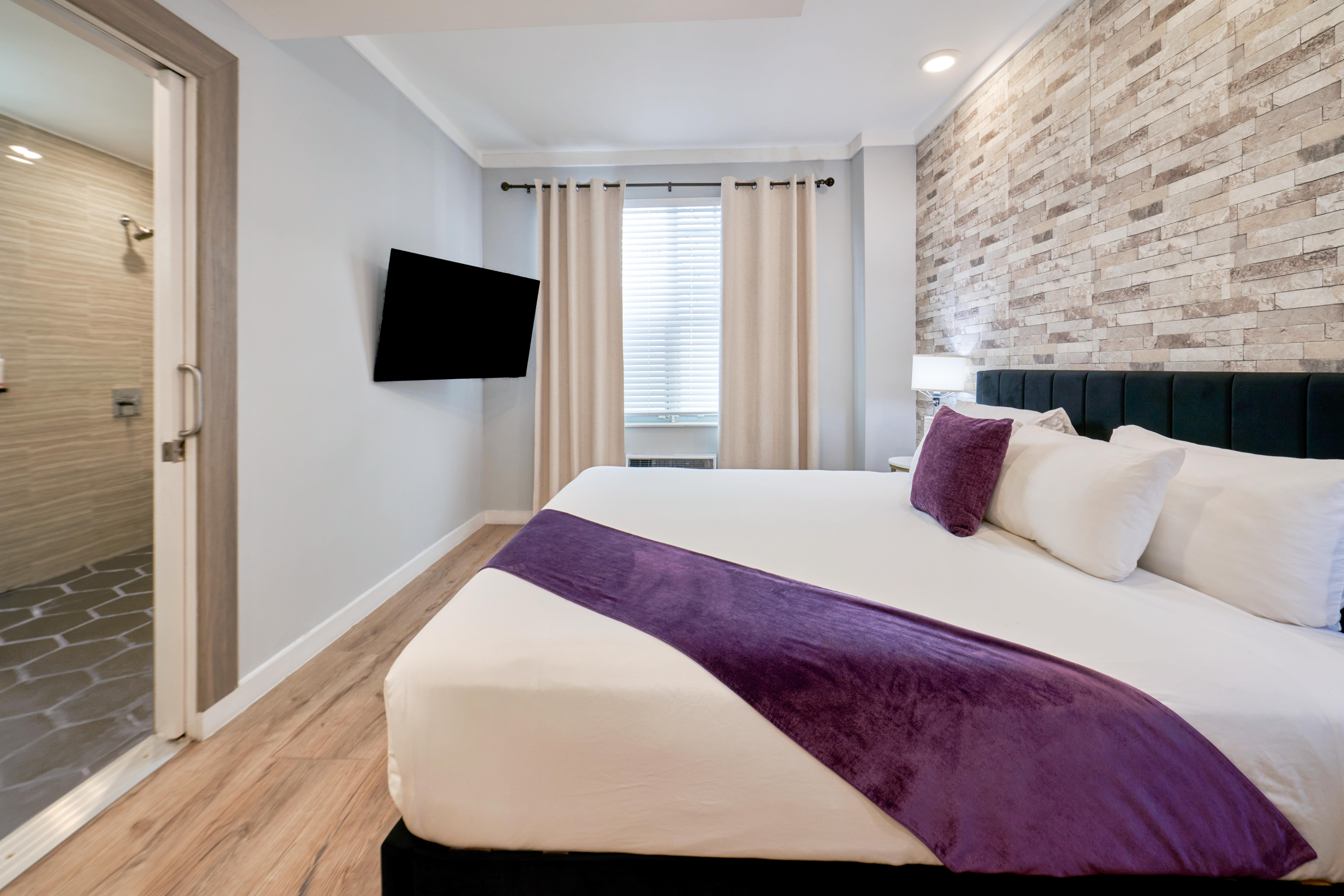 A hotel room with a large bed, a flat-screen TV mounted on the wall, and an open doorway to a bathroom with a shower.