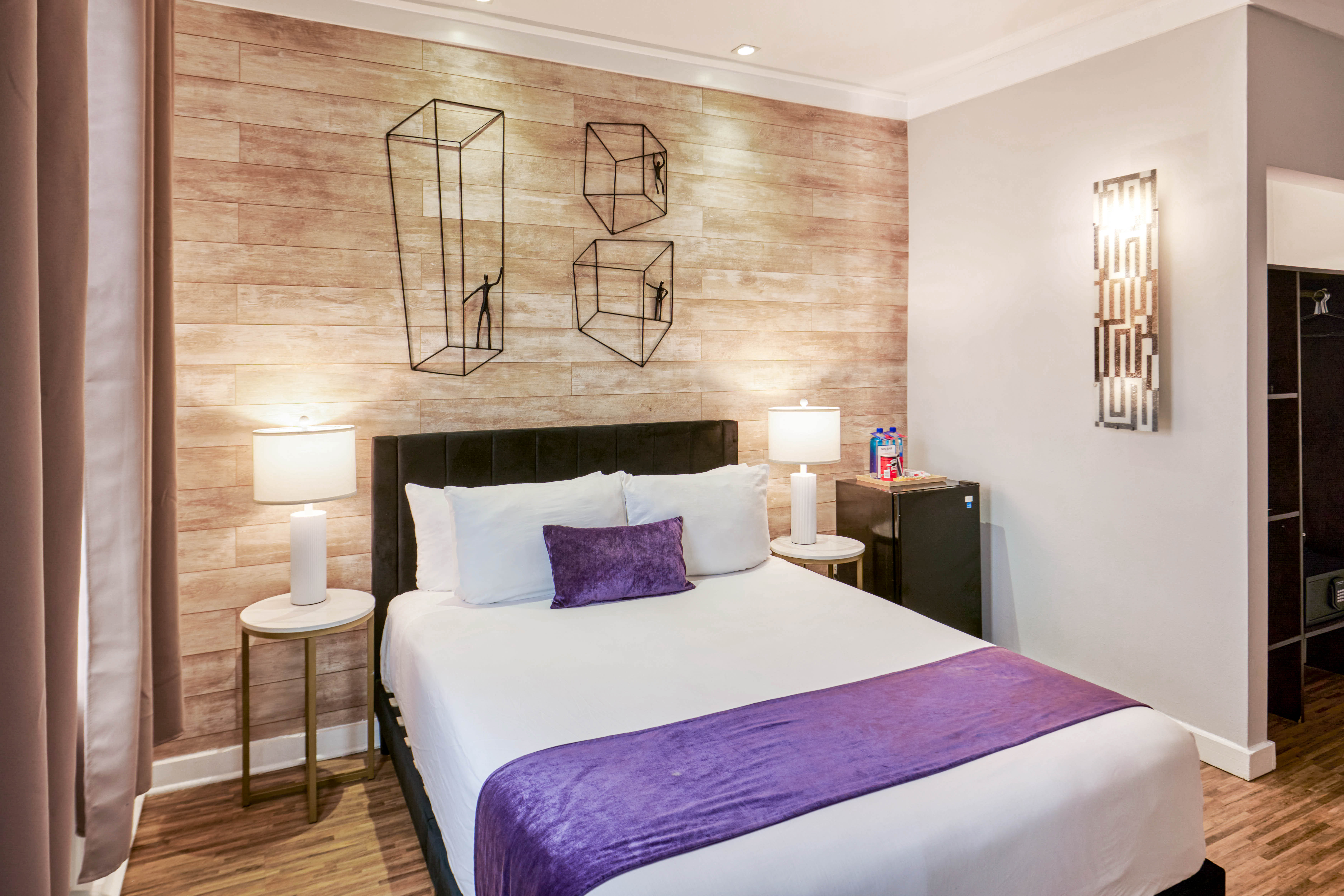 A neatly made bed with white sheets and purple accents in a modern hotel room with wood flooring and decorative wall art.